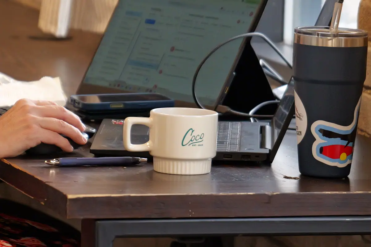 Hands at a laptop beside a branded mug and a decorated tumbler