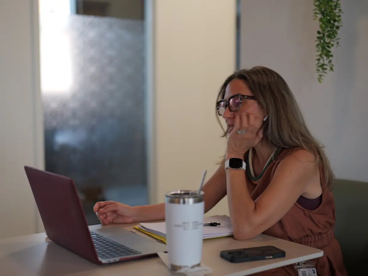 A coworking member in glasses pauses at her laptop beside a frosted-glass partition
