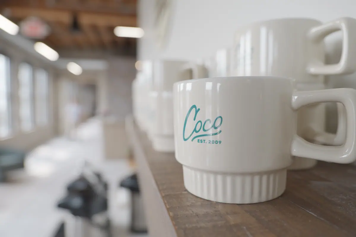 Close-up of stacked cream mugs printed with the Coco logo on a wood shelf