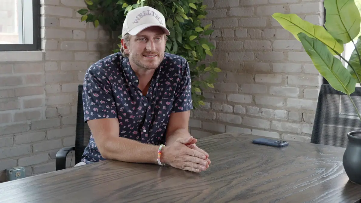 A member in a cap sits at a table during a filmed interview, brick wall and plants behind him