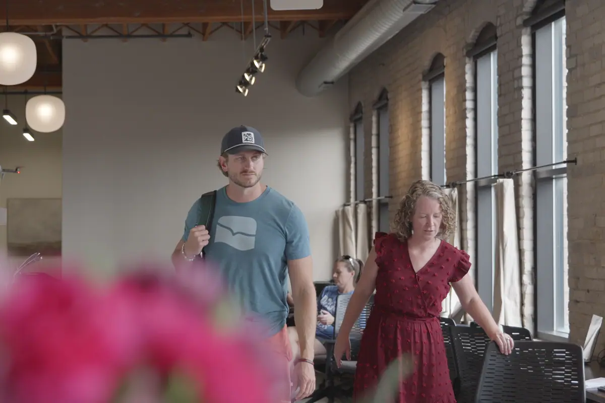 Two members walk toward camera past tall windows, pink flowers soft in the foreground